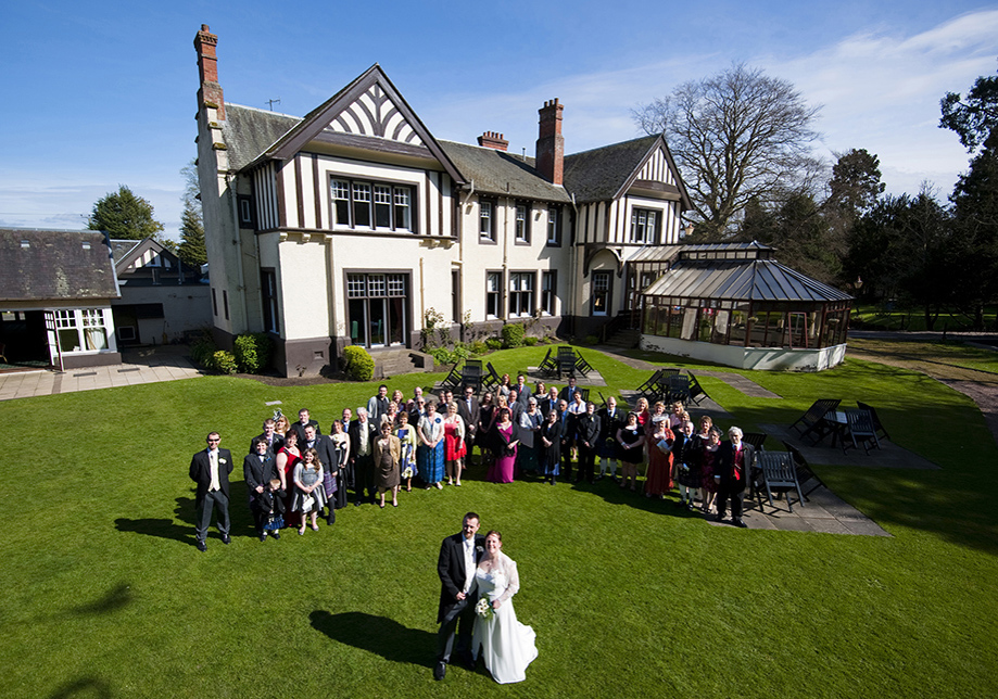 Birds eye view of wedding party on grass outside hotel with bride and groom in foreground